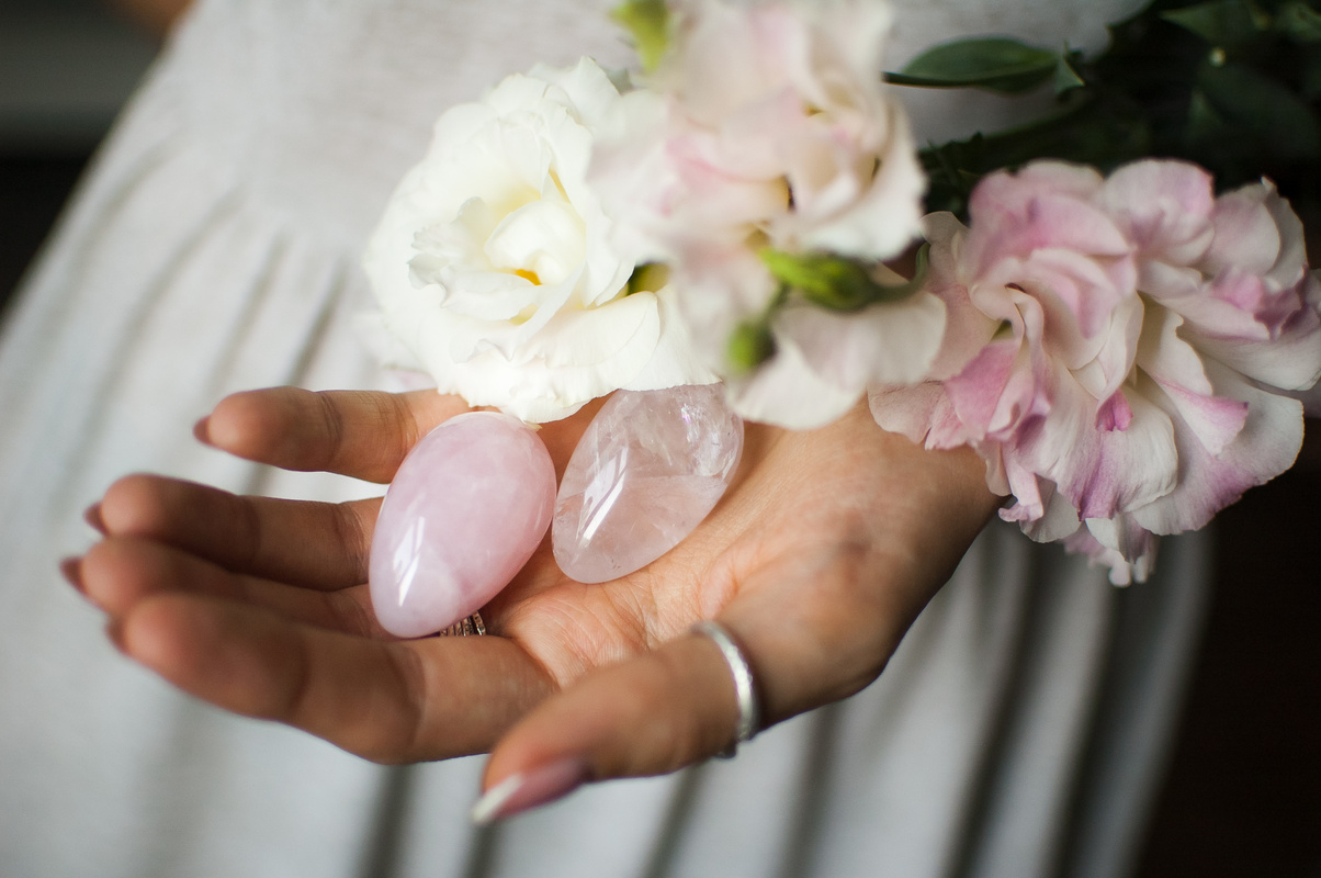 Female hands holding two yoni eggs for vumfit, imbuilding or meditation are made from pink quartz and transparent violet amethyst with white flowers indoors